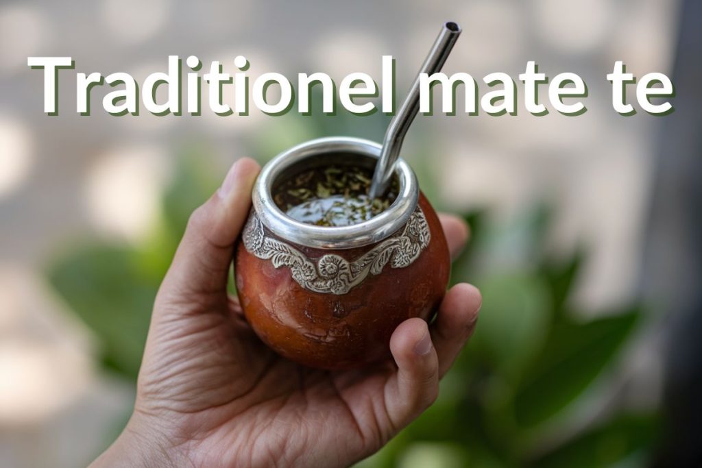 En hånd holder en klassisk kalabas matekop med en sølvkant og bombilla, fyldt med frisk yerba mate-te. Baggrunden er sløret med grønne blade i et naturligt miljø. Teksten siger "Traditionel mate te."